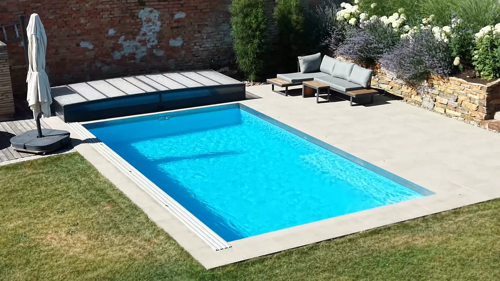 Rechteckiger Pool mit hellem Steinrand vor einem modernen Haus mit überdachter Terrasse