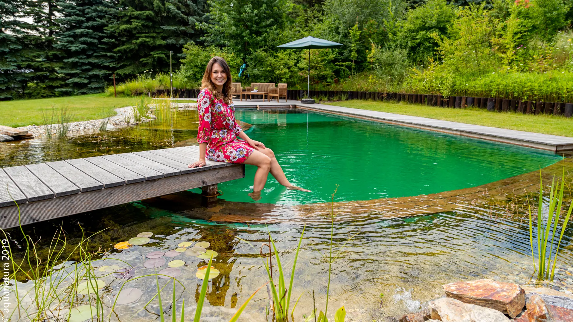 Frau auf dem Holzsteg am Schwimmteich, ringsum Seerosen und Uferstauden