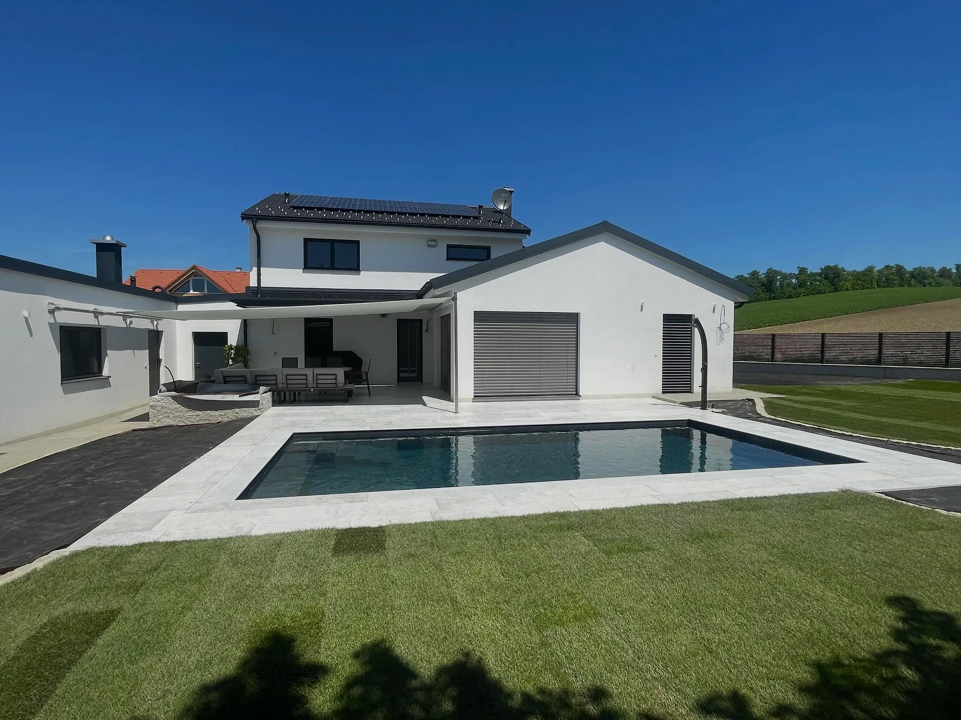 Rechteckiger Pool mit dunkler Auskleidung auf einer Terrasse aus hellen Betonplatten, dahinter Wohnhaus und Garage