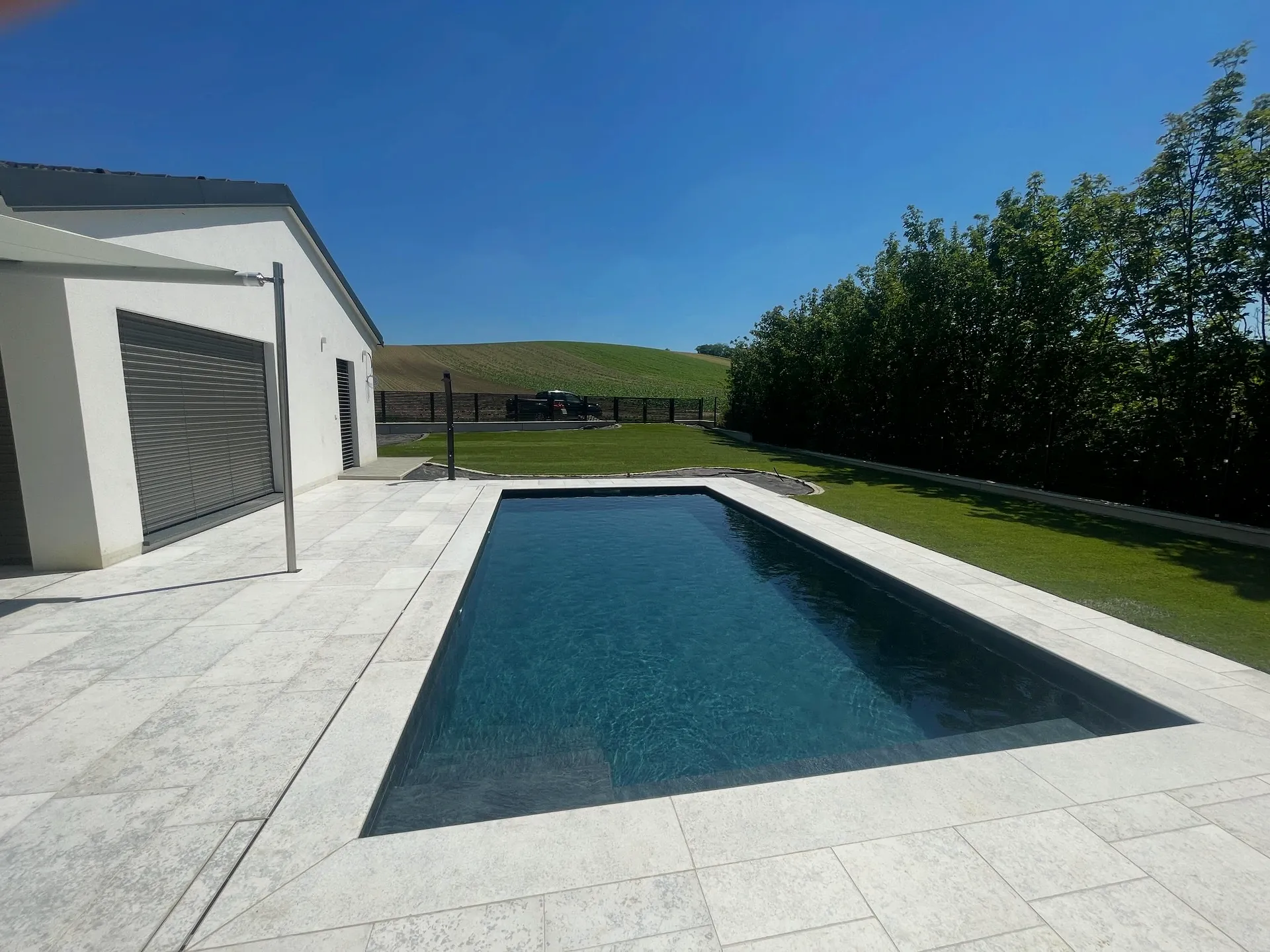 Rechteckiger Pool mit dunkler Auskleidung, umlaufender Plattenbelag und Rasen, seitlich ein flaches Nebengebäude
