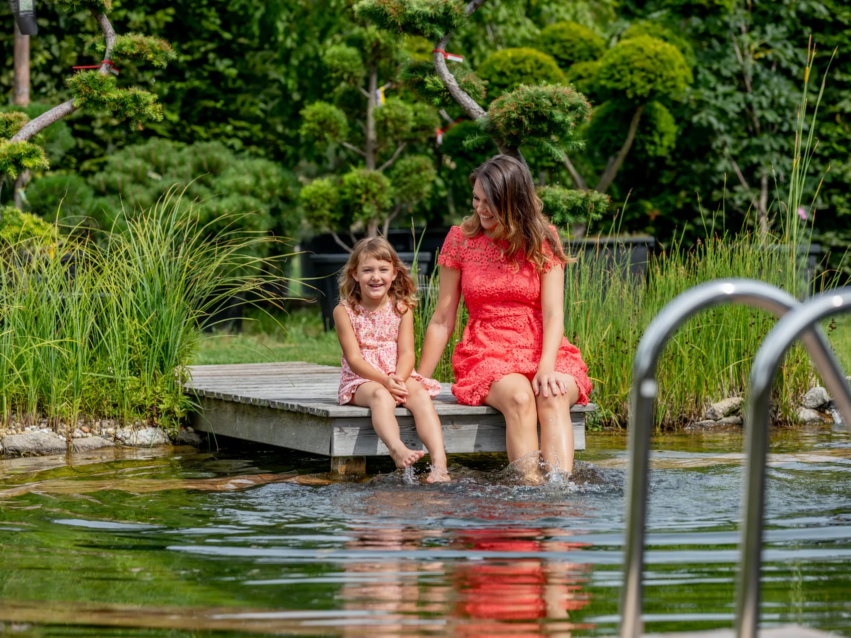 Schwimmteich mit Holzterrasse bis an die Wasserkante, dahinter zwei Wasserspeier und ein Saunahaus