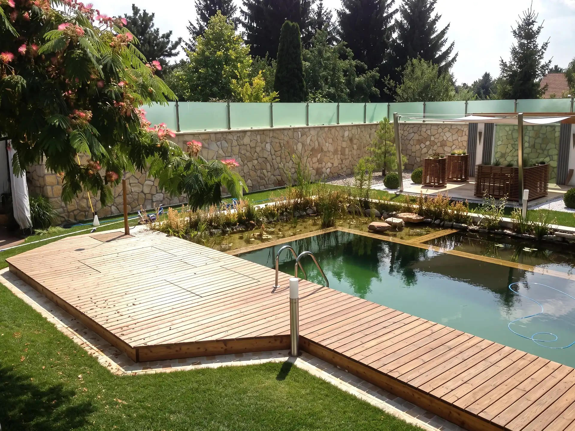 Schwimmteich mit Holzdeck und Edelstahlleiter, dahinter eine Natursteinmauer und der bepflanzte Regenerationsbereich