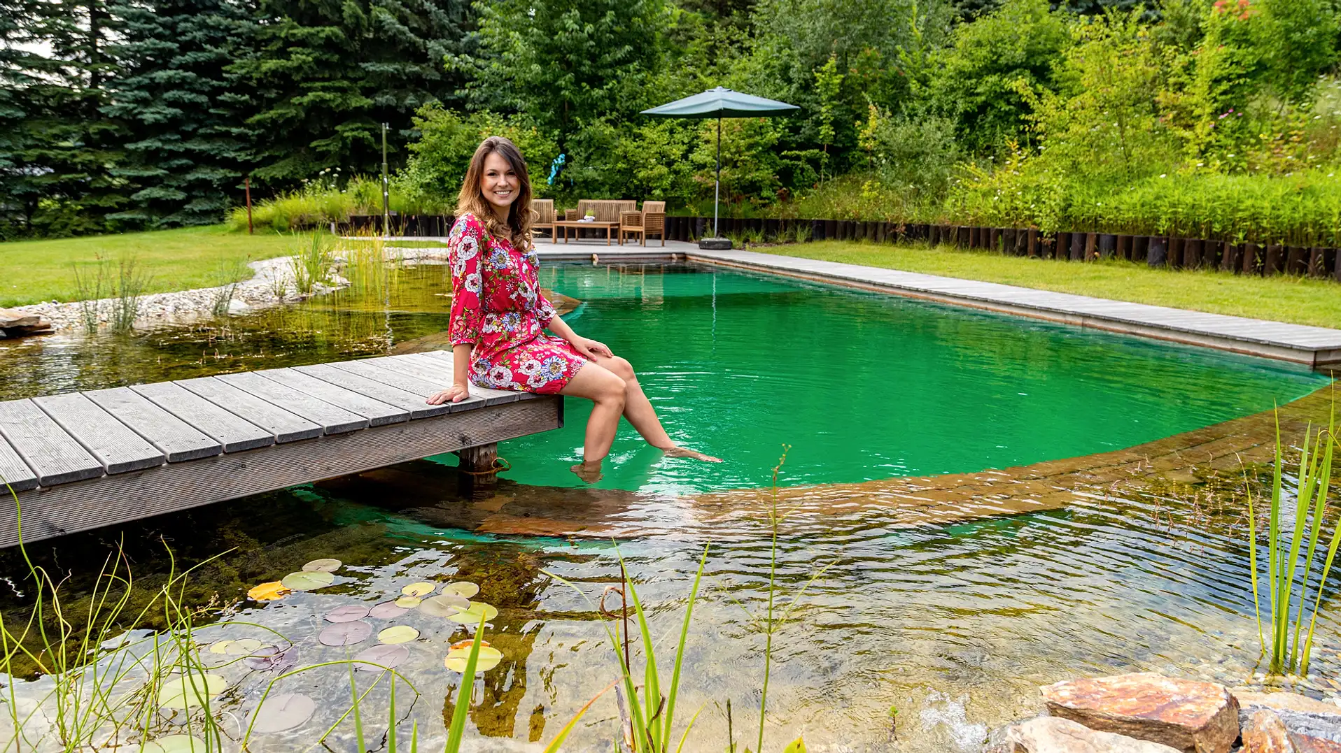 Frau sitzt am Holzsteg eines Schwimmteichs und hält die Füße ins Wasser, im Hintergrund Sitzplatz und Sonnenschirm