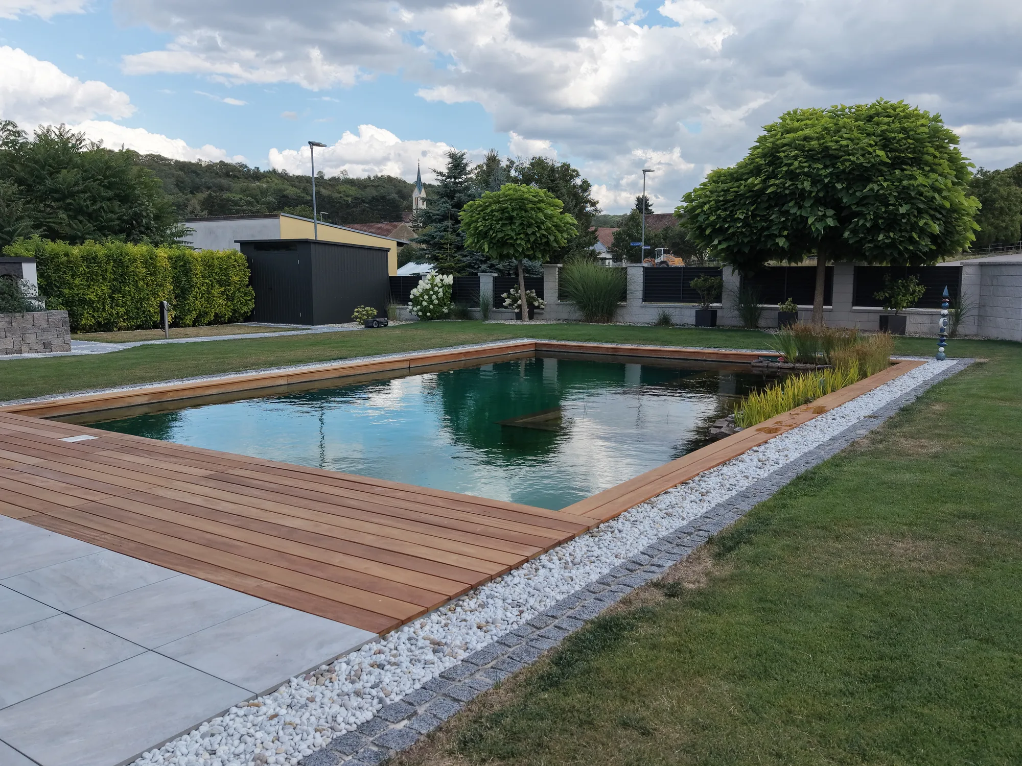 Schwimmteich mit Holzdeck, Kiesstreifen und Rasen rundherum