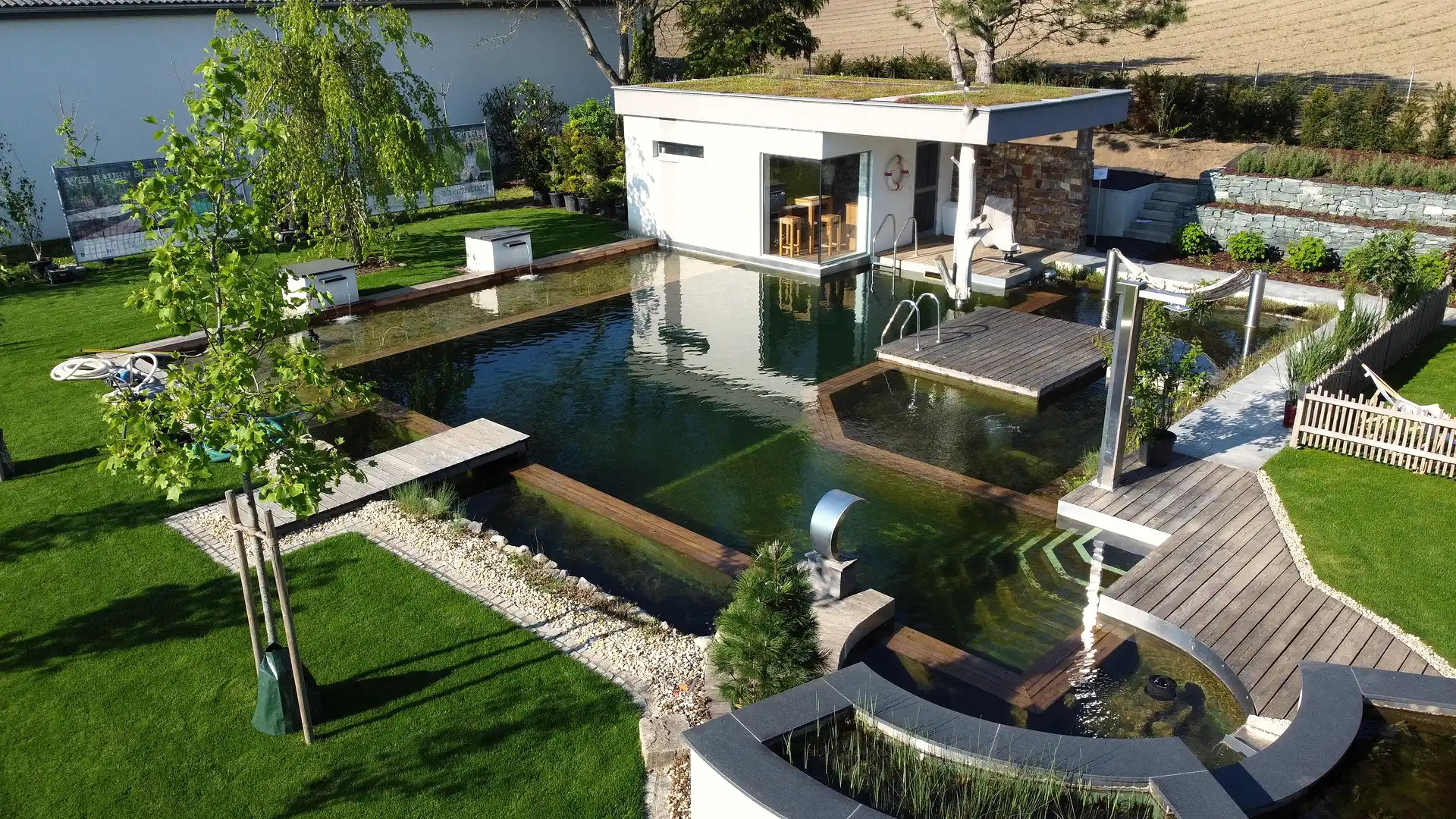 Naturpool im Schaugarten mit Wasserspiel aus Edelstahl, Poolhaus und Holzdeck